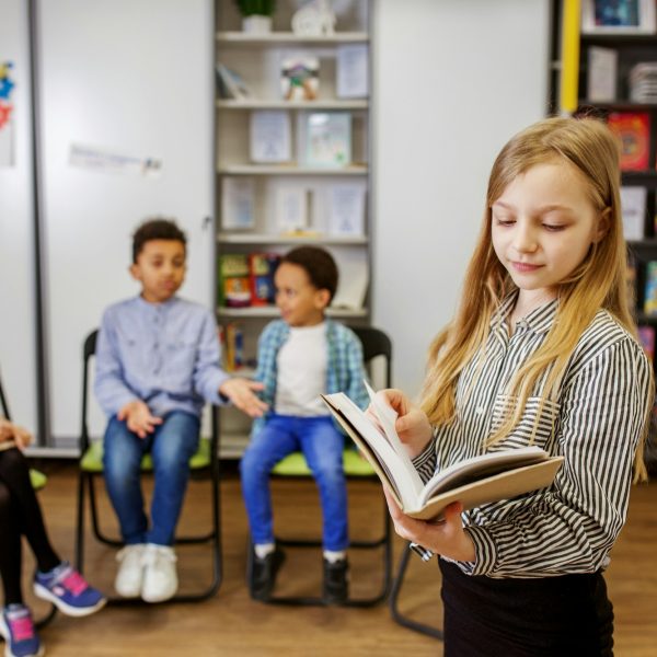 Schoolgirl standing in middle of classroom and answering question in front of classmates and teacher